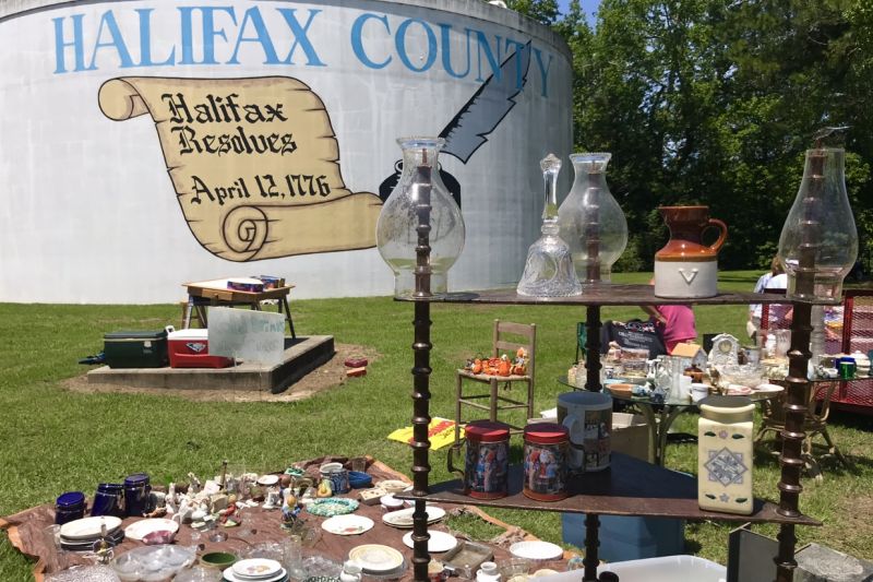 yard sale with various dishes, housewares, and furniture setup on grass in front of large water tower painted with Halifax County and a scroll showing Halifax Resolves April 12, 1776